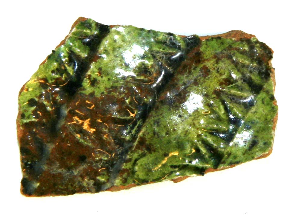 Bugskår af rødbrændt lergods, på ydersiden forsynet med indstemplede rækker af et V-formet mønster, hvorover partiel begitning af pibelersmasse, dækket af grøn blyglasur.