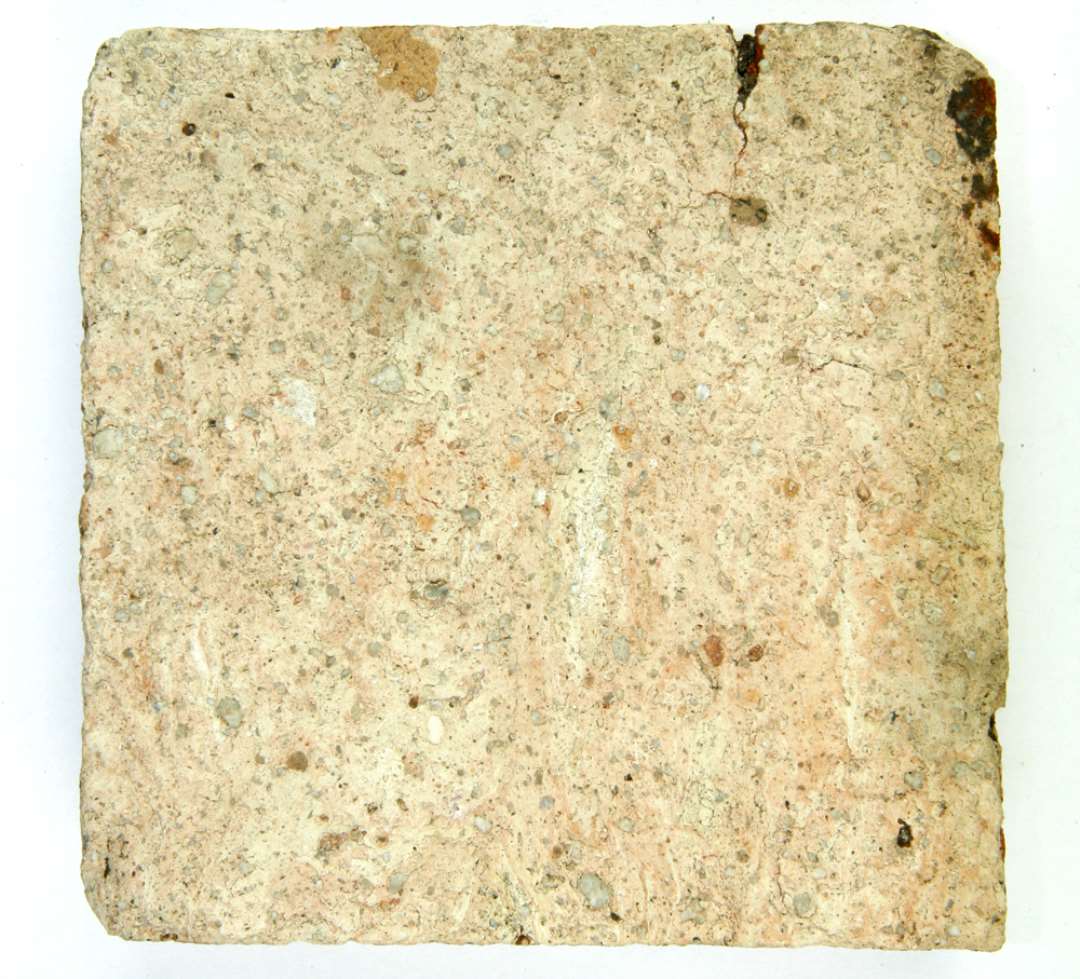 Gulvflise. Kvadratisk, af gråbrændt grusblandet lermasse. Oversiden har været dækket af nu næsten helt bortslidt klar blyglasur. Oversiden måler ca 11,3 cm i kvadrat, undersiden måler ca 10,5 cm i kvadrat, tykkelsen er 2 - 2,5 cm. 
