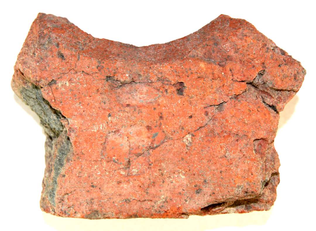 Formsten af rødbrændt tegl med sort kærne, noget fragmenteret. Stenens ene langside er plan, medens den anden er formet som en konkav indskæring, hvis ene kant tillige er affaset. Den ene kortside er ligeledes konkavt indbuet, og hjørnet mellem de to buer dannes af en ca 5 cm bred skår afskæring. Stenens anden kortside er afbrudt og lader os kun ane en begyndende konkav indskæring, der mødes i en spids vinkel med langsidens indskæring. Størst længde: ca 17 cm., størst bredde: ca 12 cm., tykkelse: knap 8 cm.