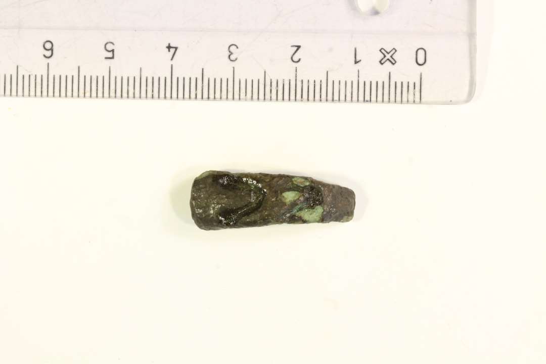 1 fragment af buet bronzearmbånd, forsynet med tre halvmåneformede, dybe indstemplinger. Markant tykkere og noget bredere i den ene ende (0,95 x 0,55 cm.), der er naturligt afsluttet, mens den modsatte ende er afbrudt (0,55 x 0,2 cm.) Bevaret længde: ca. 2,6 cm. Datering: YGJ