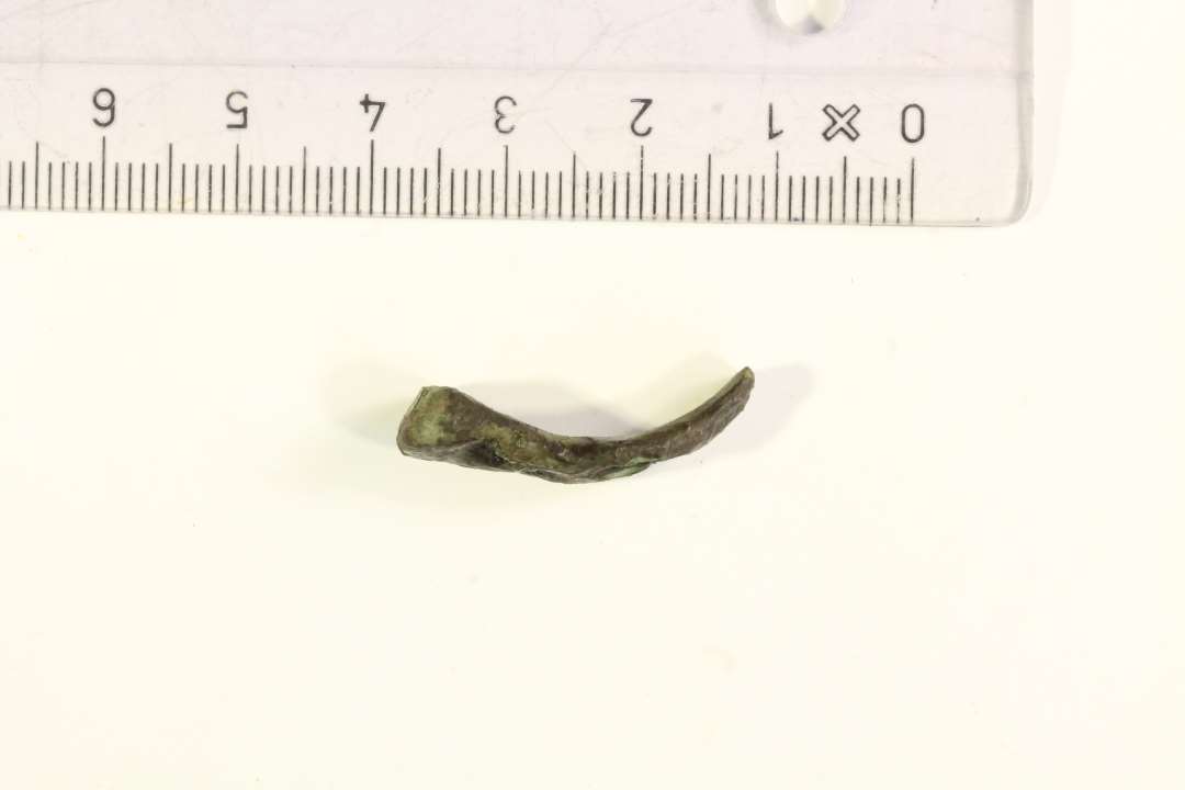 1 fragment af buet bronzearmbånd, forsynet med tre halvmåneformede, dybe indstemplinger. Markant tykkere og noget bredere i den ene ende (0,95 x 0,55 cm.), der er naturligt afsluttet, mens den modsatte ende er afbrudt (0,55 x 0,2 cm.) Bevaret længde: ca. 2,6 cm. Datering: YGJ