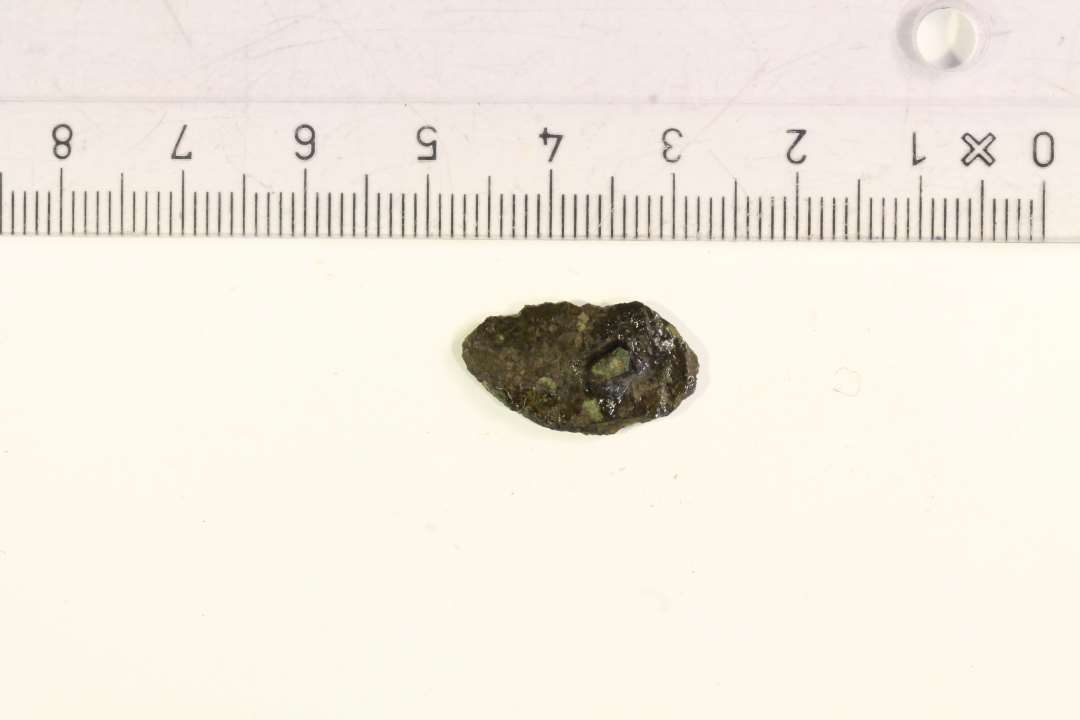 1 mindre fragment af cirkulært bronzesmykke, stærkt korroderet. Fragmenteret nålefæste eller øsken på bagsiden.  Oprindelig diameter: ca. 3 cm.