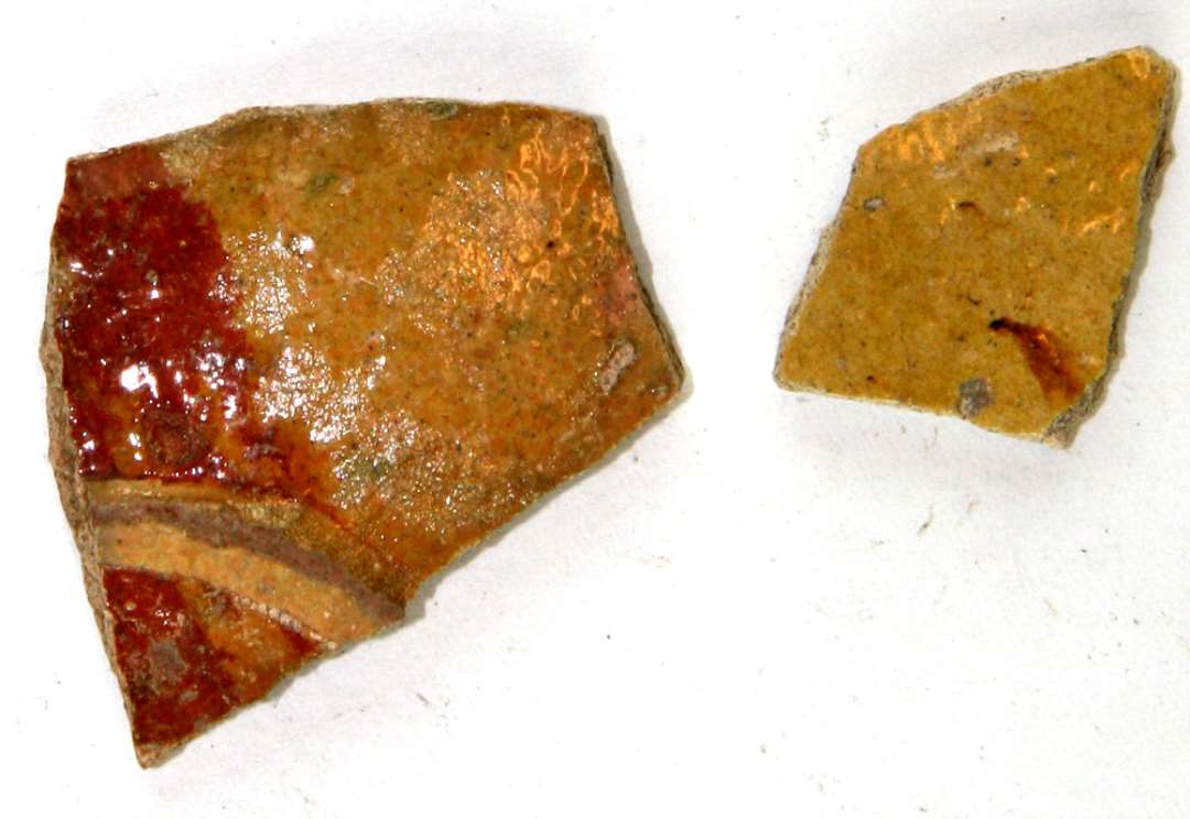 To bugskår af hårdtbrændt pibelersmasse, dækket af klar blyglasur, partiel begitning af rødler, det ene skår har tillige skarprygget pålagt lerbånd.