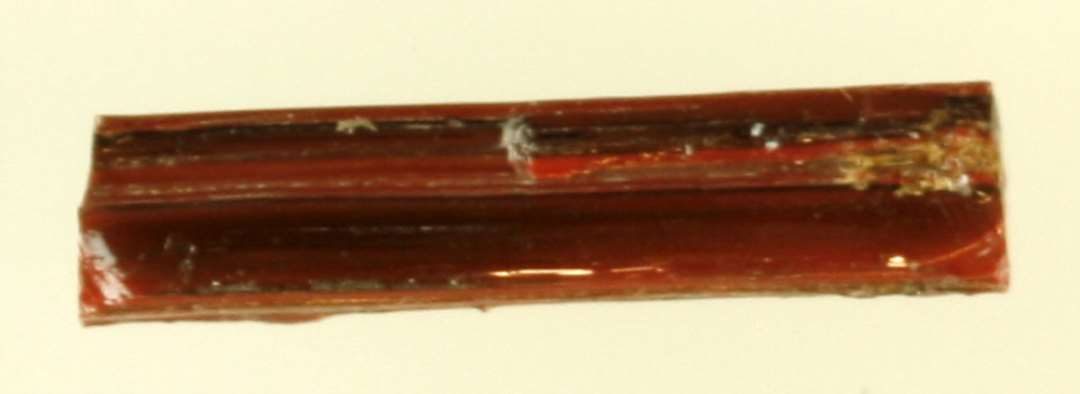 1 glasstang af rektangulær tværsnitsform, overbrudt i begge ender og af rødbrun ugennemsigtig glasmasse, hvori enkelte sorte årer. L: 1,7 cm.