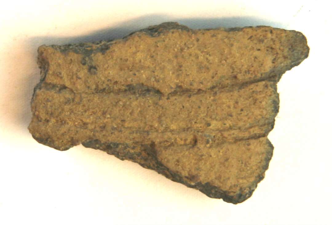 1 sideskår af brungråt lergods med dybe omkringløbende riller på ydersiden. Gr. 5.