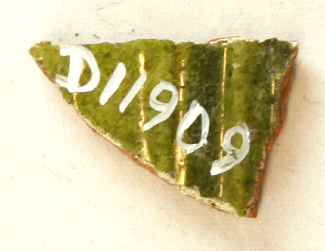 1 halsskår fra kar (kande) af rødbrændt lergods med pibelersbegitning og grønlig blyglasur på ydersiden, ujævn klar blyglasur på indersiden. Gr. 1. Flamsk