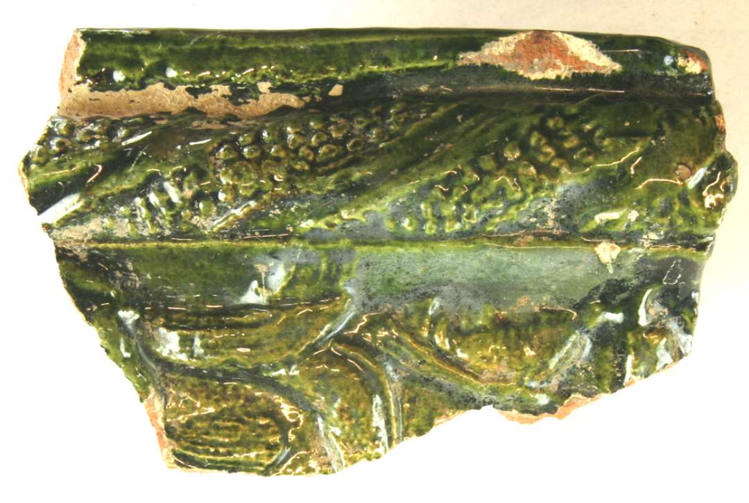 1 hjørnefragment af grønglaseret kakkel, langs hvis ene lodrette rammekant der findes en ornamenteret tovsnoet vulst. Største mål: 12 cm.