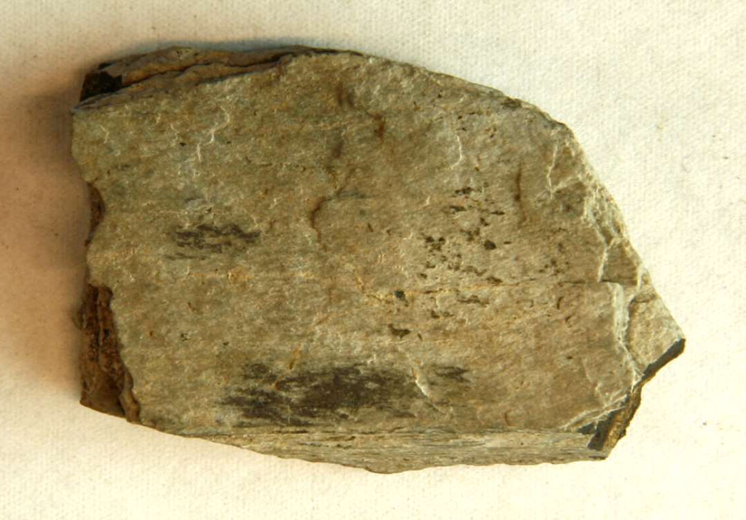1 spaltet fragment af stangformet hvæssesten af grålig skifer. Mål: 4,3 cm.