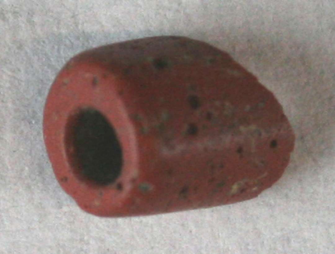 1 cylinderformet perle af rødbrunlig ugennemsigtig glasmasse med en påsmeltet omkringløbende tråd af gullig ugennemsigtig glasmasse i kanten af begge ender samt en dito i zigzagforløb på midterpartiet.