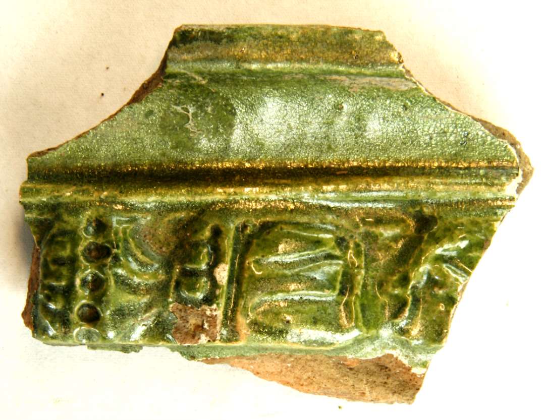 1 fragment af grønglaseret kakkel med glat hulkehlsrammekant og rest af ornamenteret søjle i hovedfeltet. Største mål: 7,5 cm.