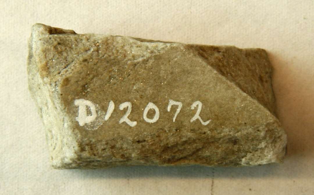 1 aflangt fragment antagelig af stangformet hvæssesten af grålig, ildskørnet sandsten. L: 3,5 cm.