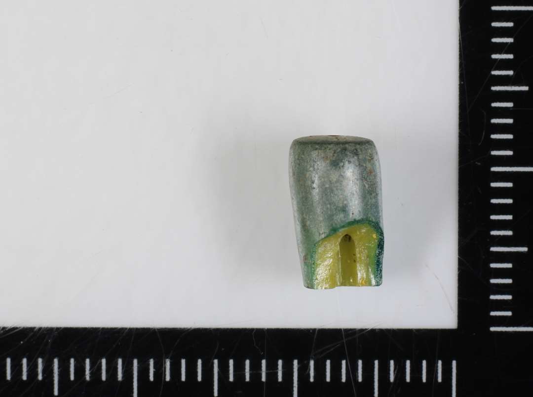 1 fragmenteret, delvis smeltet, cylinderformet perle af gullig ugennemsigtig glasmasse omgivet af en tynd kappe af grønlig ditto. 9 mm.
Teknisk er der tale om en trukken glasrørsperle.