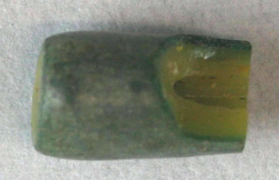 1 fragmenteret, delvis smeltet, cylinderformet perle af gullig ugennemsigtig glasmasse omgivet af en tynd kappe af grønlig ditto. 9 mm.
Teknisk er der tale om en trukken glasrørsperle.
