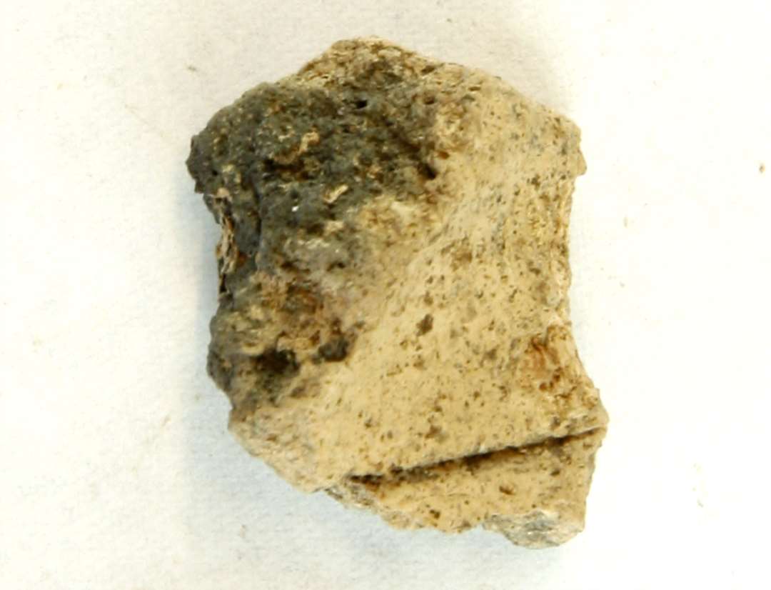 1 fragment fra ophængningshullet på discosformet vævevægt af brungrå brændt lermasse. Mål: 3 cm.