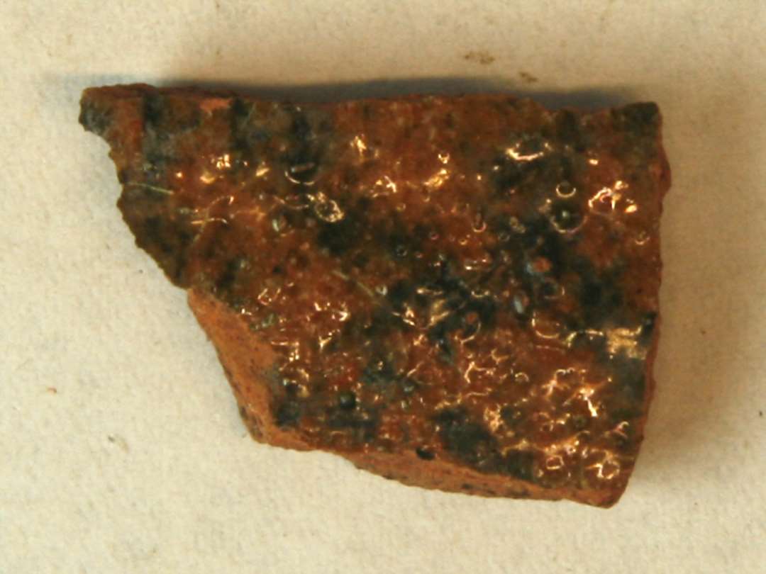 1 sideskår af rødbrændt lergods med klar blyglasur spættet med sortbrun jernoksyd på ydersiden, pletter af klar blyglasur på indersiden. Gr. 1.