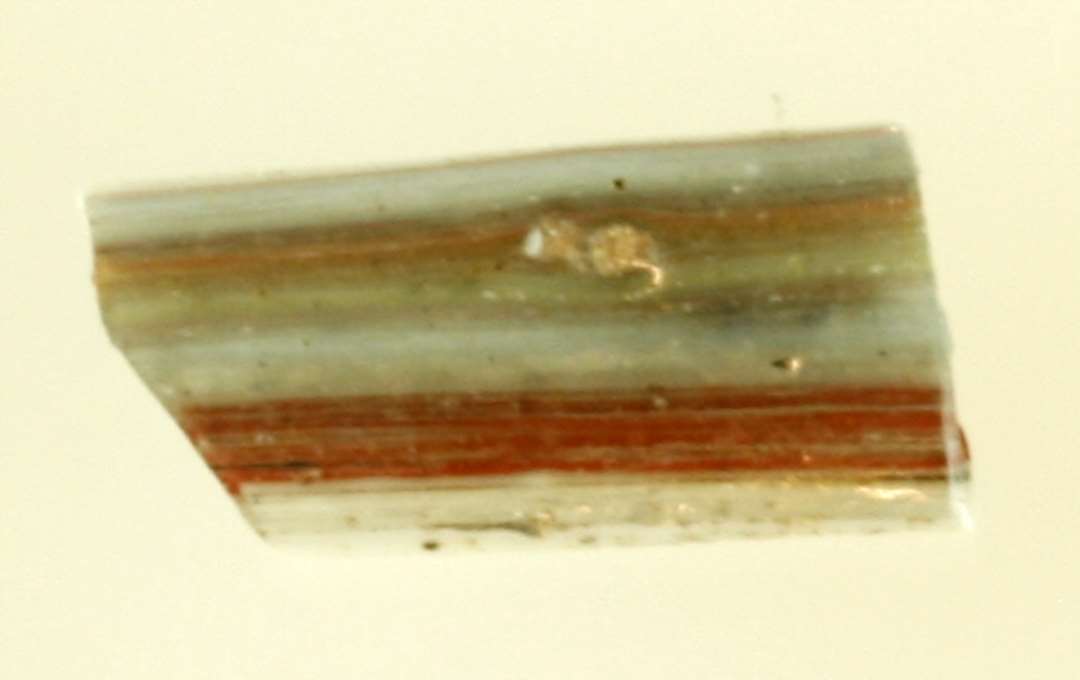1 fragment af flad bændelformet glasstang bestående af tråde af afvekslende rød og hvid ugennemsigtig glasmasse tildels sammenholdt af et tyndt lag blålig gennemsigtig glasmasse på stangens ene side.