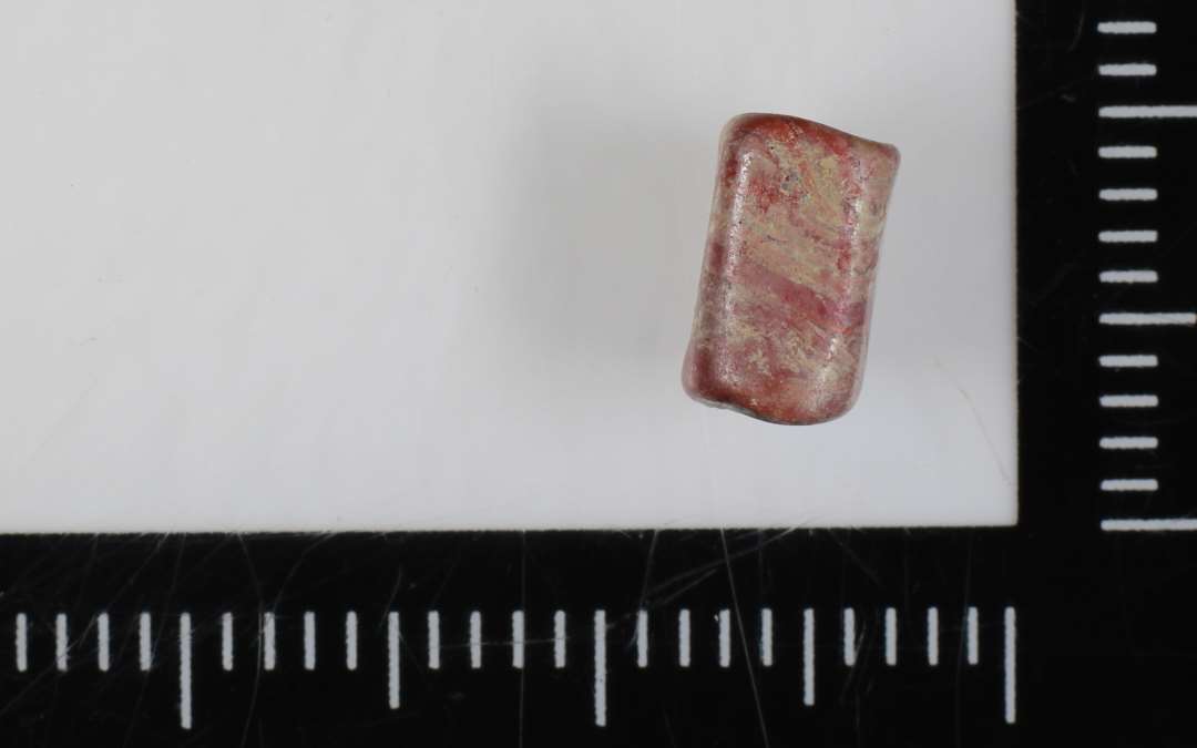 1 cylinderformet perle med kvadratisk tværsnisform med afrundede kanter af rødbrunlig ugennemsigtig glasmasse.  L: 0,7 cm.