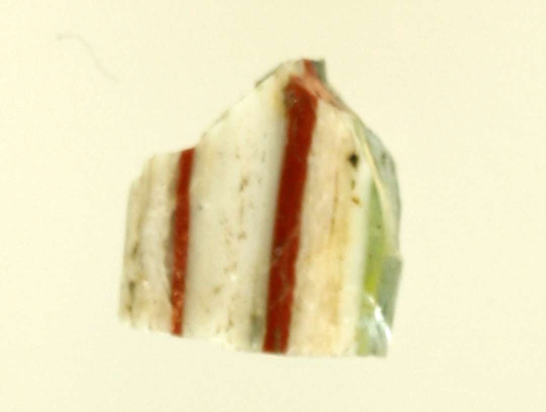 Brudstykke af stang af glas, den ene halvdel af grånligt glas, den anden af mælkehvidt med indlagte røde tråde.  L: 0,7 cm.