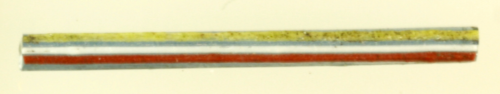 1 stang af blåligt gennemsigtig glas af rektangulær tværsnitsform. På den ene sideflade er fastsmeltet tre tynde tråde af ugennemsigtig rødbrun, hvid og gullig glasmasse. L: 3,1 cm.