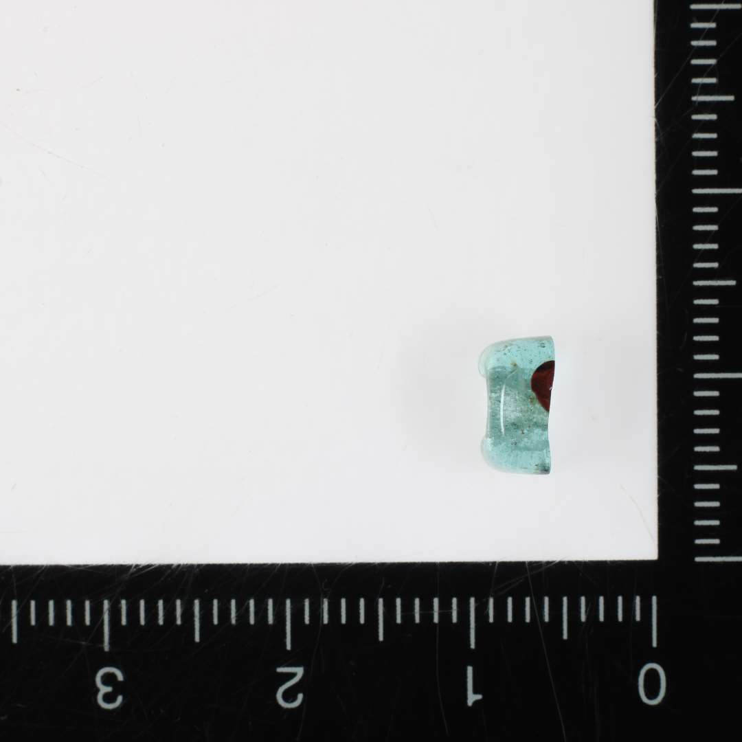 1 fragment af perle af gråligt, gennemsigtigt glas med spor af indsmeltede pletter af rødbrunlig, ugennemsigtig glasmasse. Perlen synes at have haft kvadratisk tværsnitsform med afrundede hjørner og endekanter. 6 mm.