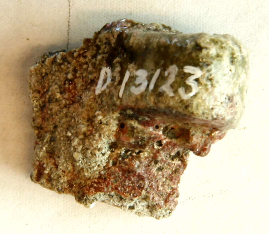 1 sideskår fra smeltedigel af gråbrændt porøst lergods med glasuragtig sintringsskorpe på ydersiden, hvor der findes en vandret fremstikkende, afrundet gribetap. Mål: 3,3 cm.