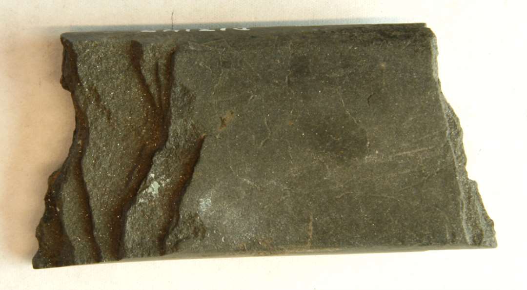 1 fragment af hvæssesten af glatsleben finkornet, mørkegrå bjergart af flad stangform overbrudt i begge ender i skrå uregelmæssige brud. L: 7,4 cm. Br: 3,8 cm. Tyk: 1,4 cm.