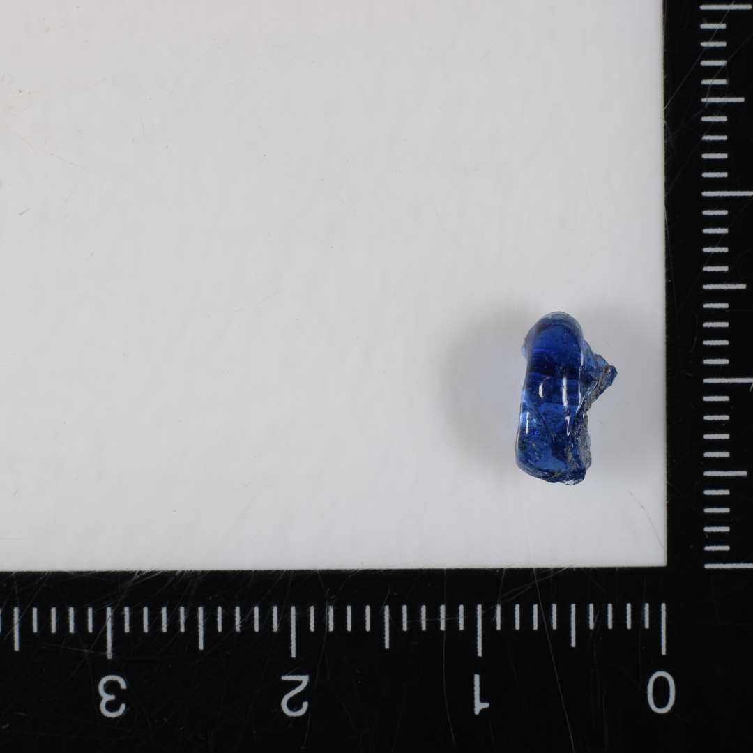 1 fragment af perle af blålig, gennemsigtig glasmasse af afrundet ringform med ujævne smeltningsspor på den ene sidekant. 9 mm.