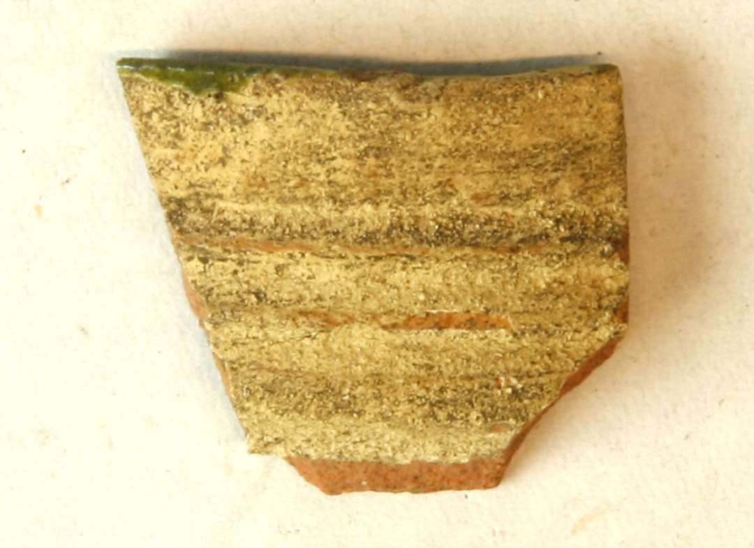 1 randskår fra stjærtpotte af rødbrændt lergods med pibelers begitning på ydersiden samt på indersiden, der er dækket af grønlig blyglasur. Gr. 1.
