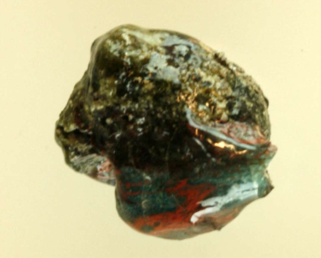 1 klump smeltet, delvis sintret glas af størrelse som et stort solbær og af delvis gennemsigtigt blågrønligt glas med årer af rødbrunlig ugennemsigtig glasmasse.
