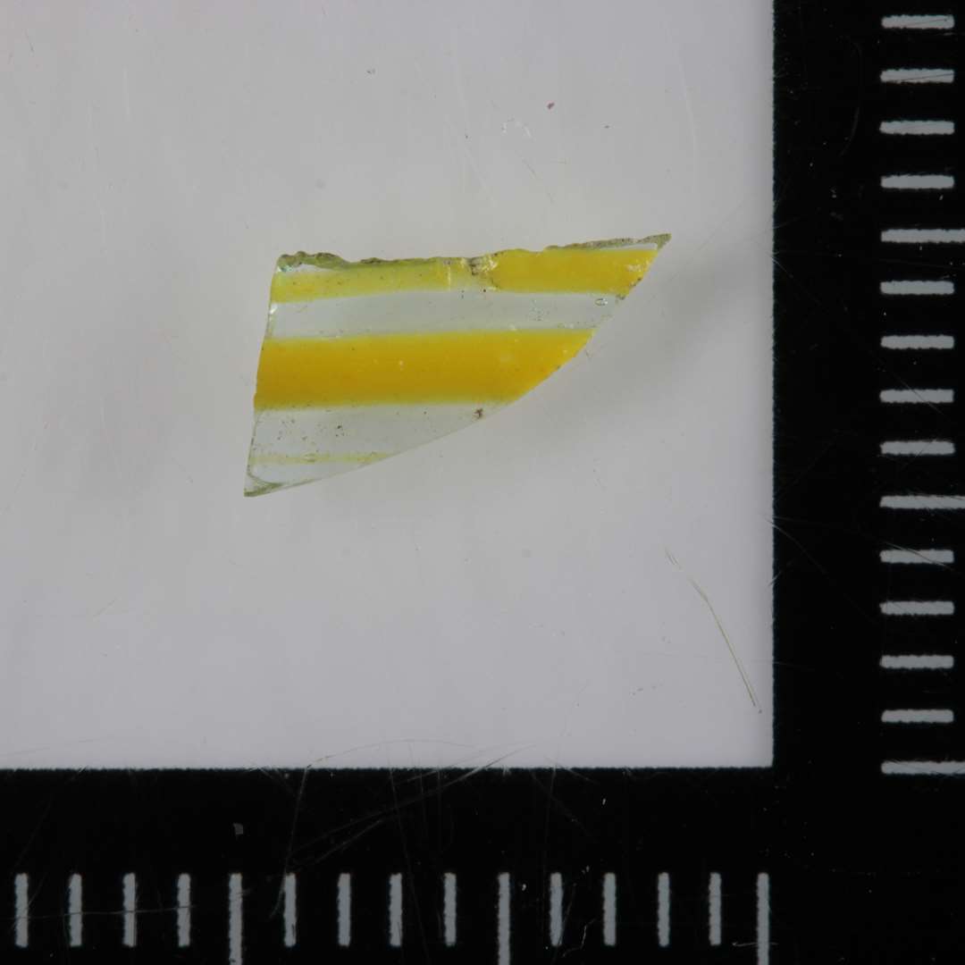 1 ubetydeligt sideskår fra kar(bæger) af klart gennemsigtigt glas med påsmeltede flade trådribber af gult ugennemsigtigt glas på ydersiden.