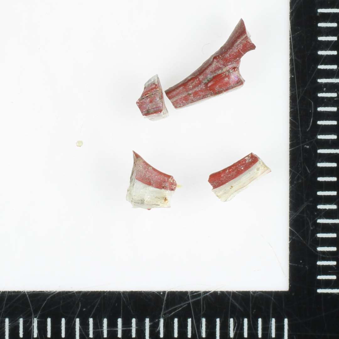 12 fragmenter af glastråde, heraf 8 af flad, tildels bændelagtig form, 4 af tynd trådform, alle dannede af sammensmeltede tråde af ugennemsigtig rødbrunlig og hvidgår glasmasse.