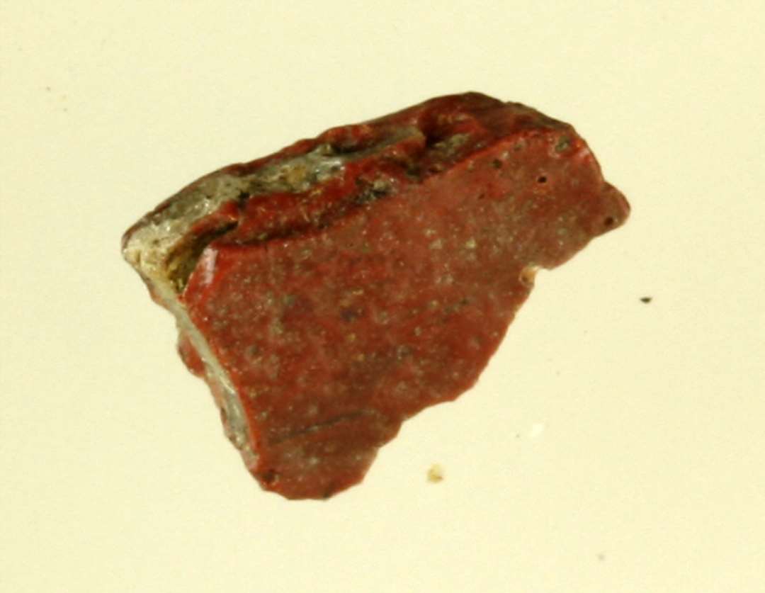 1 flad fragmentstump af rødbrunlig, ugennemsigtig glasmasse i hvis midte sidder indkapslet et lag af ugennemsigtig grågrumset glasmasse.
