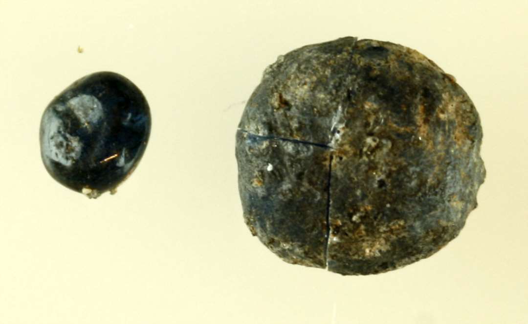 2 fladovale kugler af smeltet til sintret blåligt gennemsigtigt glas med iltet overflade, den ene af størrelse som et nipsenålshovede, den anden som en stor ært.