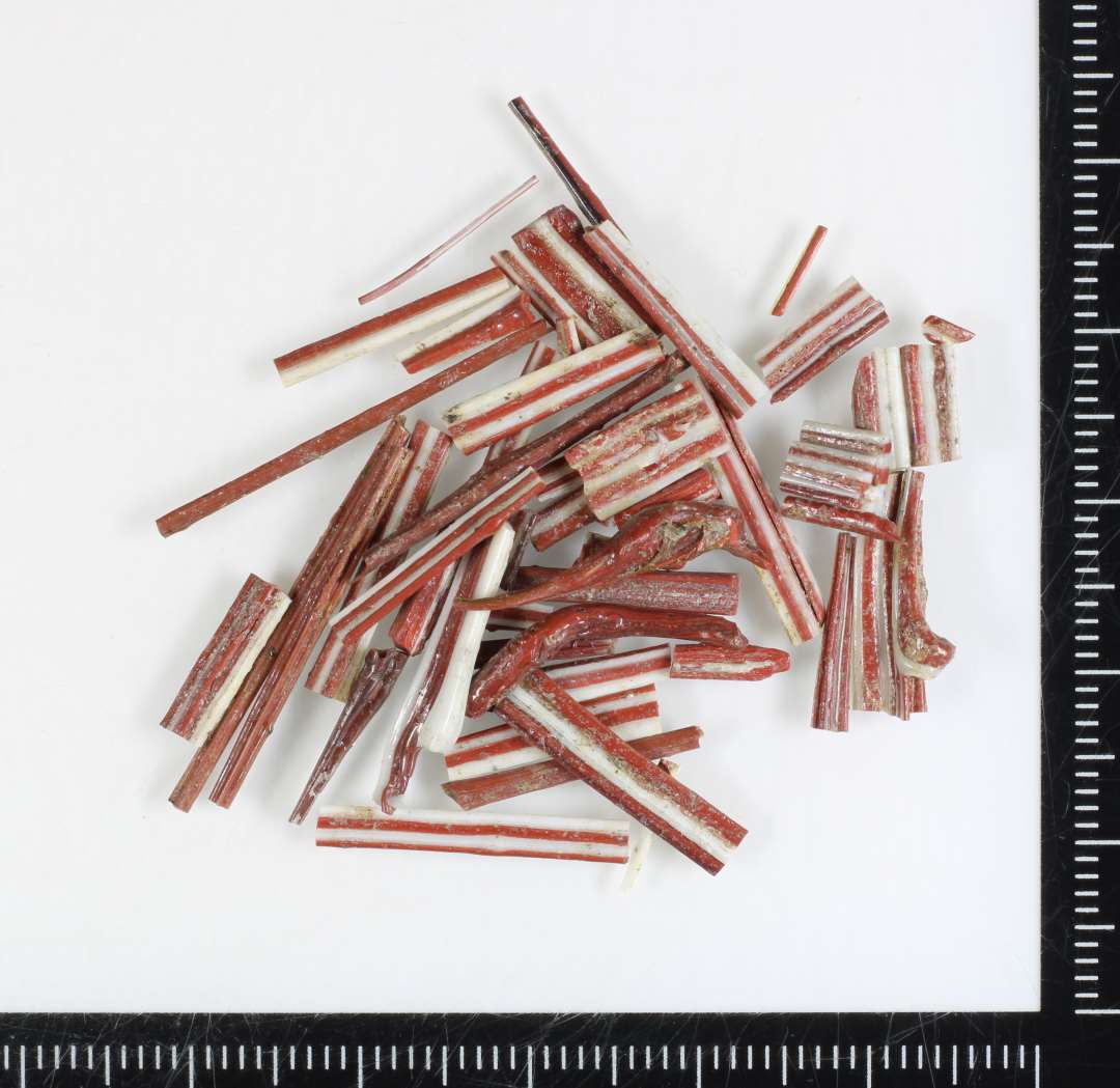 En samling fragmenter af varierende smallere og bredere, flad bændelagtigt stangform bestående af sammensmeltede tråde af ugennemsigtig hvidgrå og rødbrunlig glasmasse.
