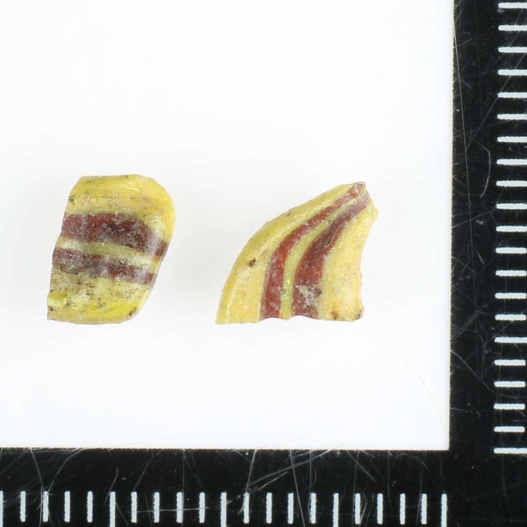 En samling fragmentstumper af flad bændelagtig stangform bestående af sammensmeltede tråde af ugennemsigtig gullig og rødbrunlig glasmasse.  Sammenhørende med D13484.