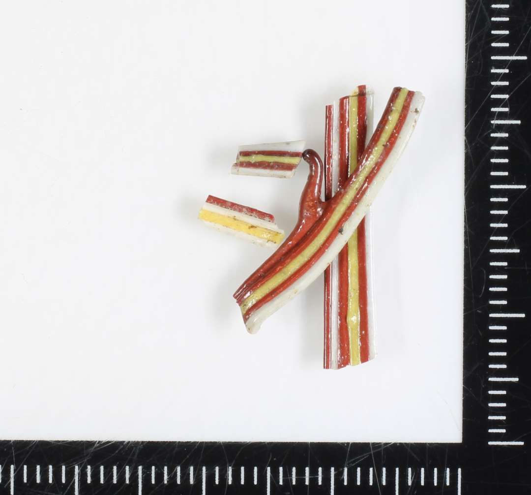 En samling fragmentstunper af flad bændelagtig stangform bestående af sammensmeltede tråde af ugennemsigtig hvidgrå, rødbrunlig og gullig glasmasse.
