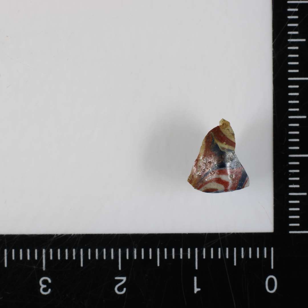 1 fragment af perle af dobbeltkonisk form af gennemsigtig blålig glasmasse, hvori på ydersiden er indsmeltet tråde af ugennemsigtig hvid og rødbrunlig glasmasse i et udflydende mønster. 10 mm.
