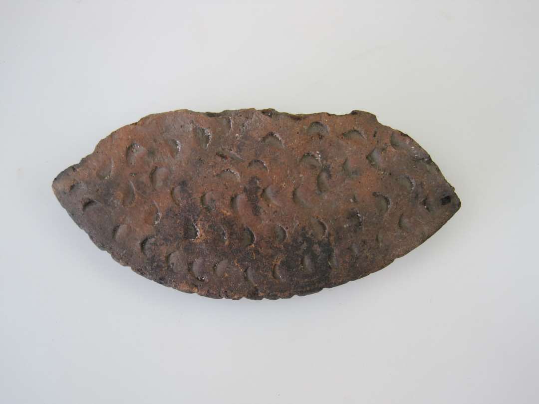 Fragment af låg af teglgods. Med primitiv, indstemplet ornamentik. Største mål: 11,7 cm.