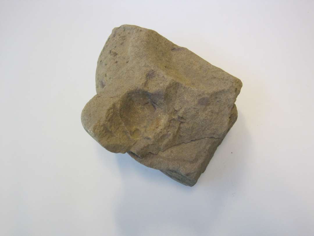 Fragment af flad, cylindrisk løber, hugget i porøs sandsten, som er lagdelt i gulliggråt og violet. største mål er 19x12 cm., højden er 11,4 cm. På oversiden er ved stenens midte en antydning af en lav, tragtformet åbning til indhældning af kornet bevaret. Fra denne forløber en ophøjet ribbe ud til stenens rand. I ribbens kant er en hugget fordybning, hvori et løst drejehåndtag kan have været placerert. Stnens underside og side er blankslidt.