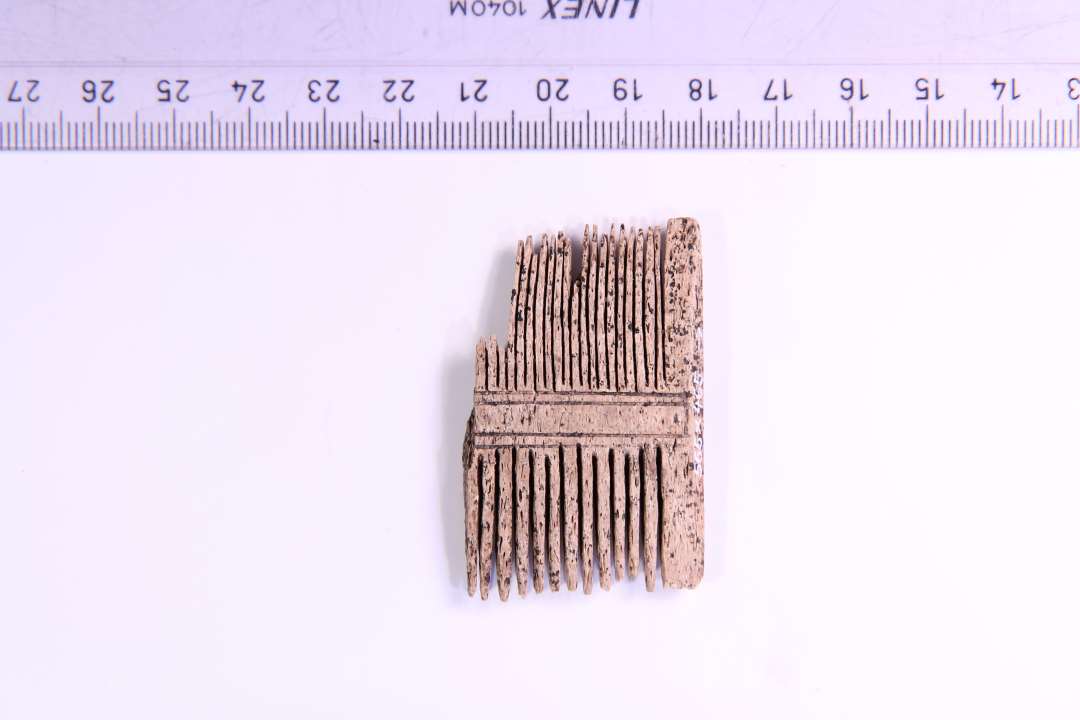 Fragment af dobbeltkam af ben i et stykke. Længde 3,1 cm. Højde: 4,9 cm. Med både store og små tænder. Udsmykket med to dobbeltlinier.