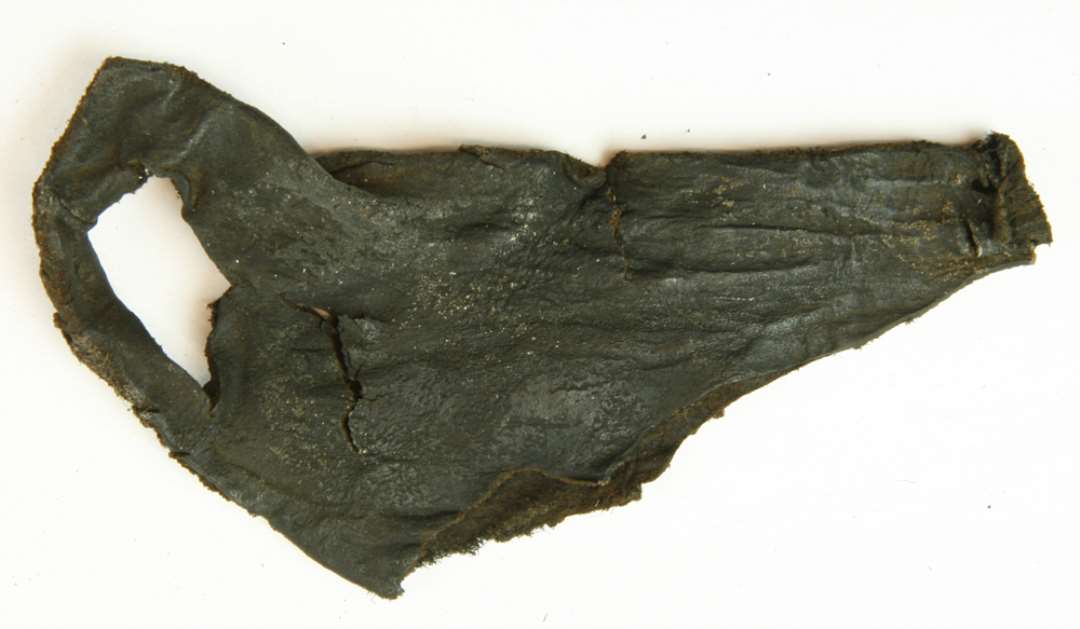 1 forrevet og forskåret læderfragment af nærmest tresidig form  ca. 6,5 -14 cm. med en større skævt fortrukket ca. 3,5 cm. lang, snøreåbning langs den ene rand.