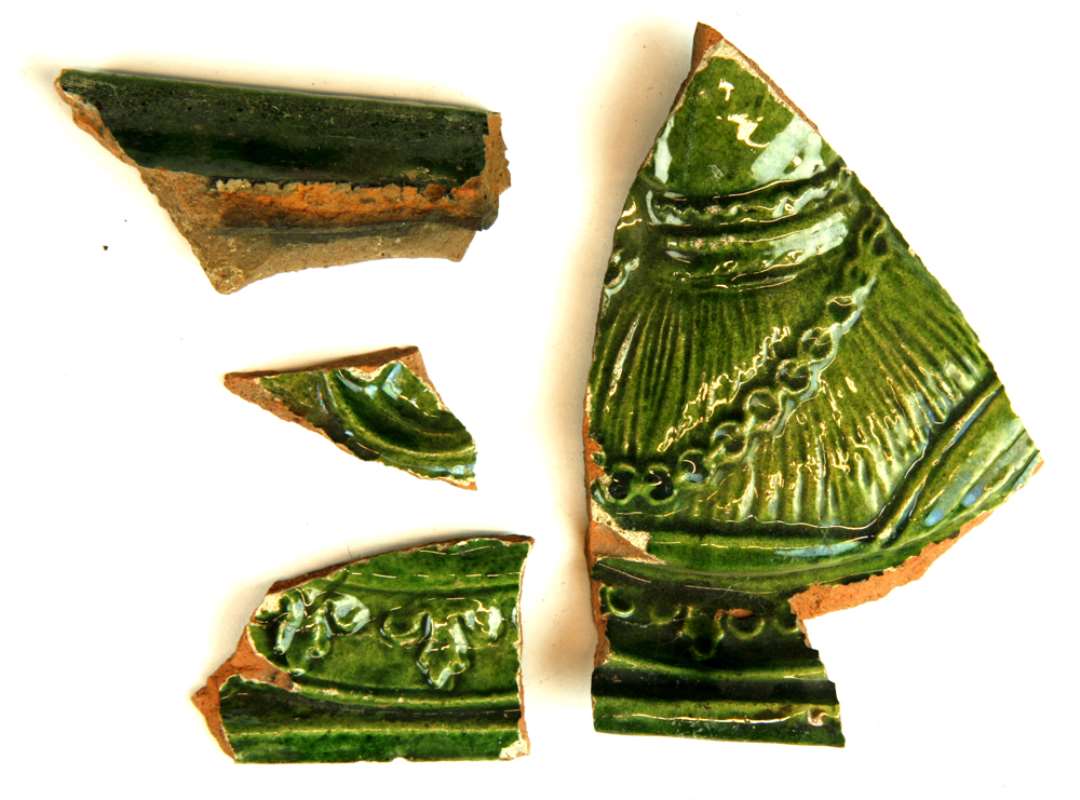 4 fragmenter fra ramme og midterfelt af grønglaserede, dekorerede ovnkakler. Et af skårene fremviser et udsnit af brystpartiet på en person i renæssancedragt med kraftig halskæde.