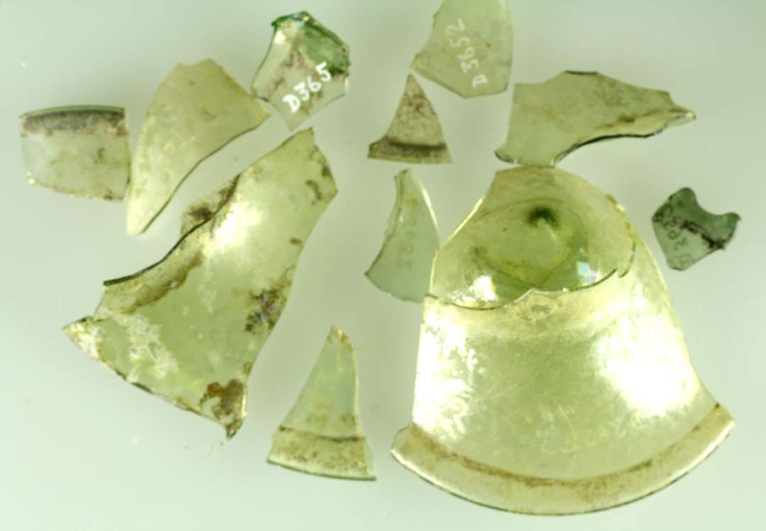 23 større og mindre fragmenter af opadhvælvede bunde og fødder med hul vulst langs ståfladen, af klart, let grønligt glas, stangenglas.
