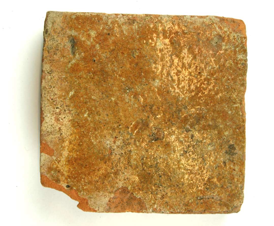 1 let fragmenteret gulvflise af rødbrændt lermasse med tynd pibelersbegitning og klar blyglasur på oversiden.  Største mål 13,3 X 13,6 cm. Tykkelse 3,5 cm.