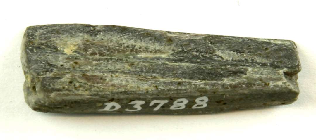 1 fragment af en slibesten af grå skifer. Stykket er 3 cm. langt, har et trekantet tværsnit og er afbrudt i begge ender. Største mål: 3,8 cm.