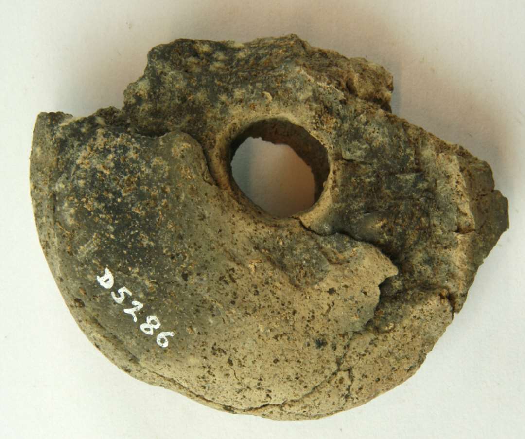 1 fragmenteret vævevægt af discoform af gråbrunlig brændt lermasse, diam. ca. 8,2 cm.
