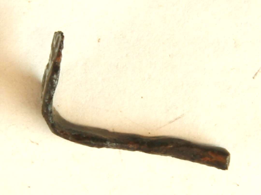 jernfragment af trind stangform, vinkelret ombøjet i den ene ende, der er udhamret i flad form. L : 2 cm.