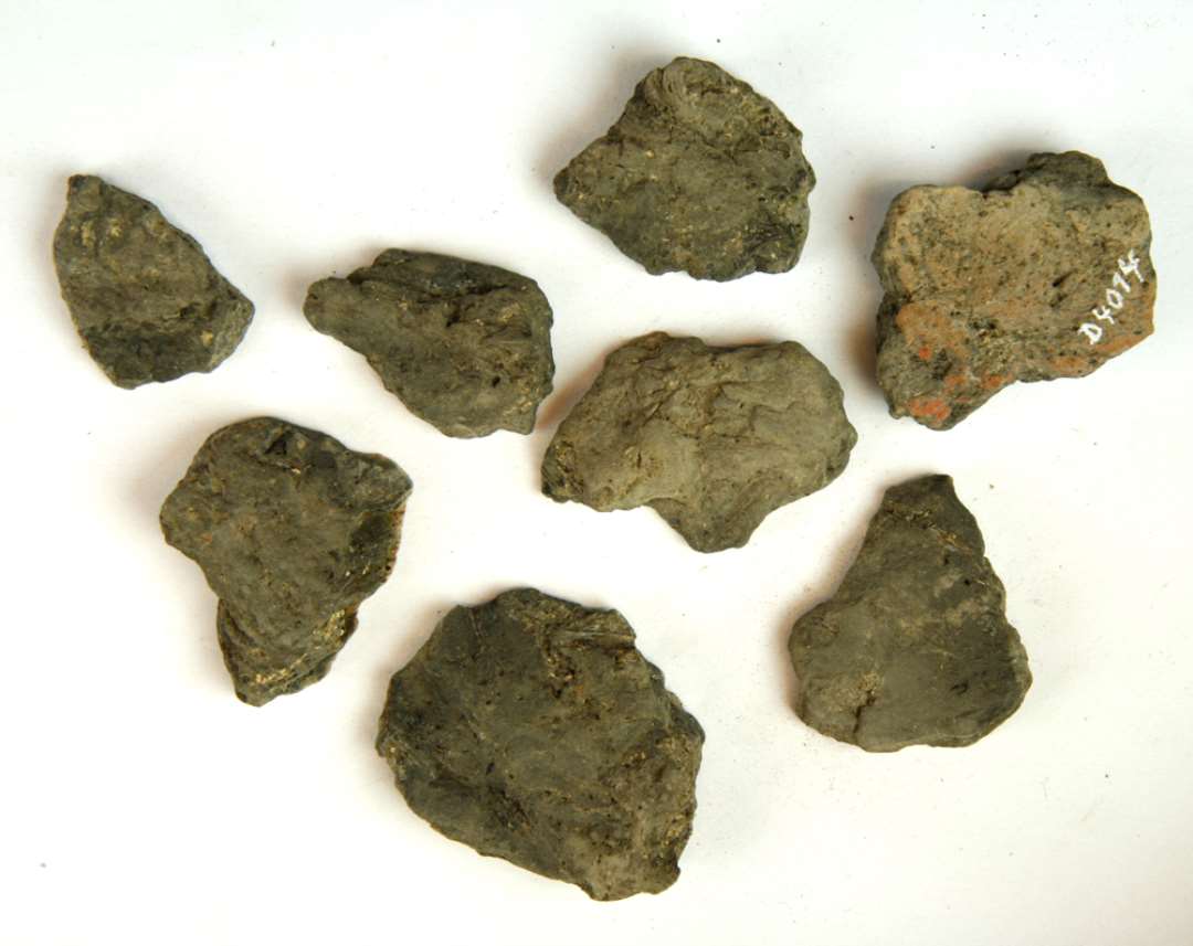 8 fragmenter af gråbrunlig brændt lermasse med mørkegrå kærne og delvis plan yderside. Støbeform? Mål: ca 3-5 cm.