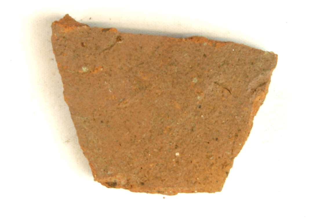 Topskår fra sparebøsse af rødbrændt lergods med en enkelt ubetydelig plet af klar blyglasur på ydersiden. På skåret ses svage drejeriller, og rester af den flade tut på toppen af sparebøssen. Største mål: 4,5 cm.