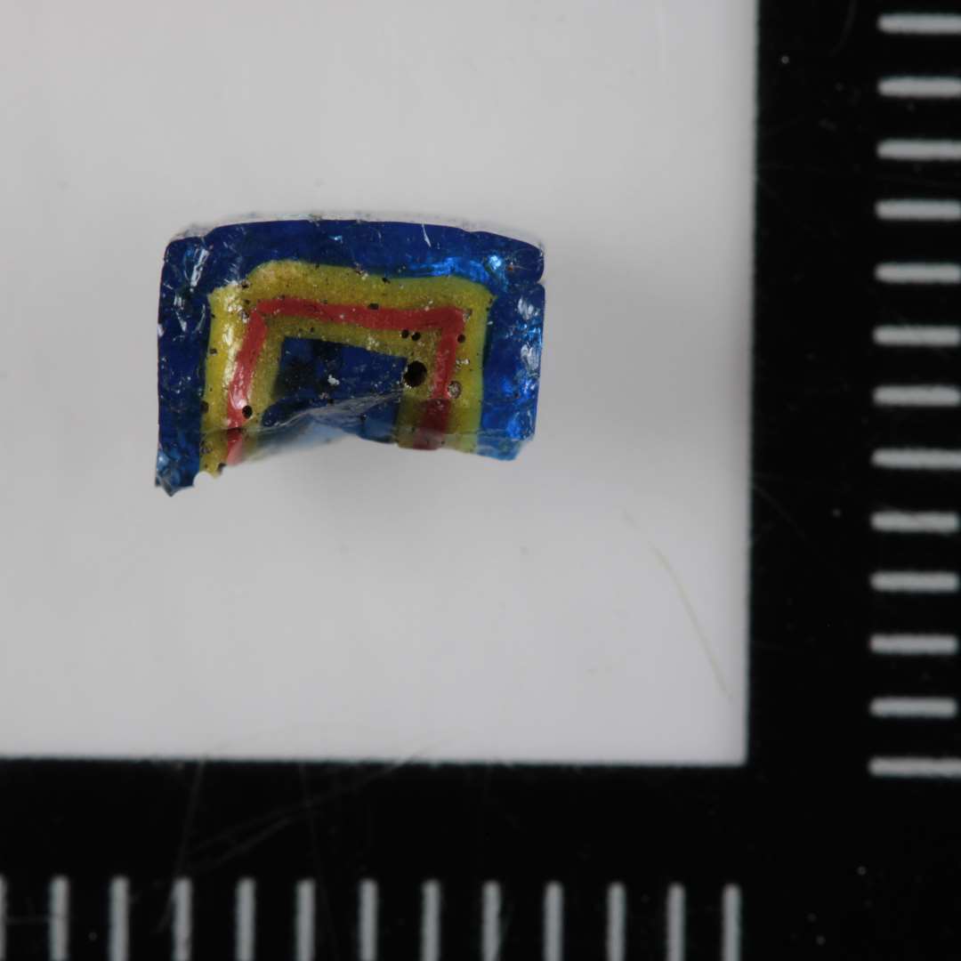 1 fragment af perle ( ? ) af firkantet form bestående af lag af skiftevis blåt, gult og rødt glas.